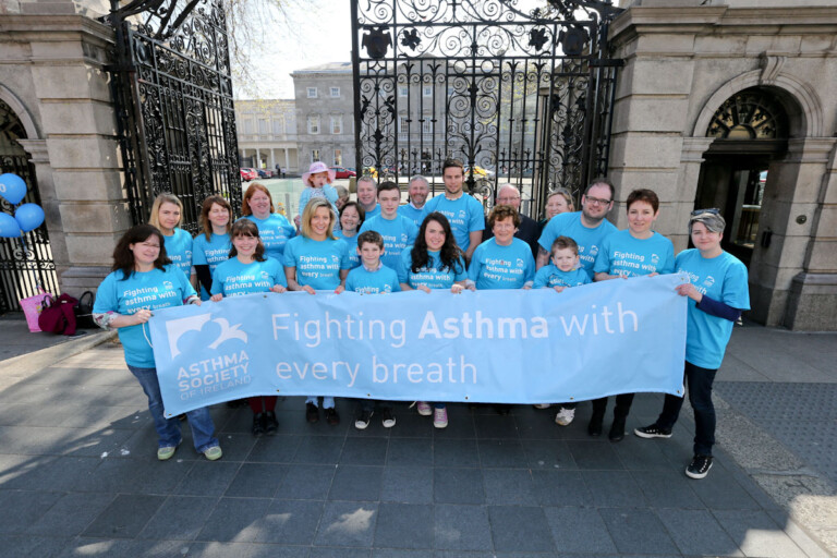 A group of people wearing matching blue T-shirts hold a banner reading "Fighting Asthma with every breath" in front of ornate iron gates, conveying unity and advocacy.