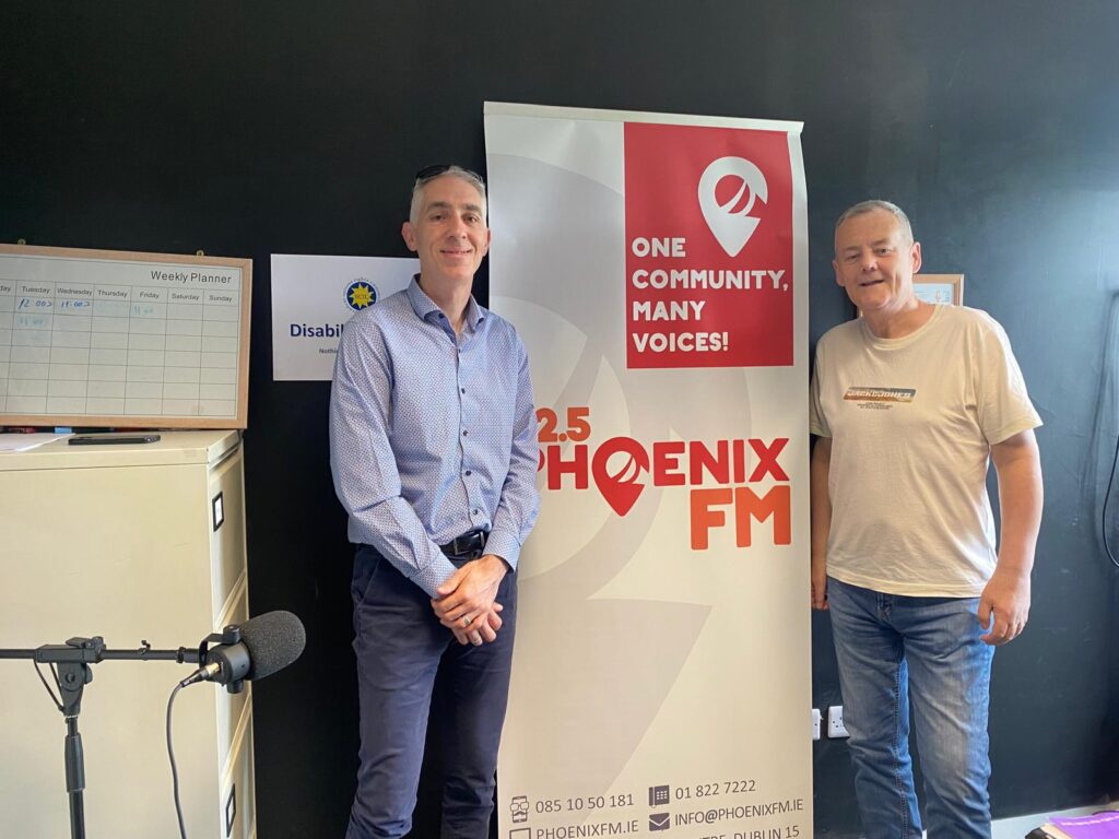 Two men stand smiling near a "92.5 Phoenix FM" banner with the slogan "One Community, Many Voices." A microphone and a planner are visible nearby.