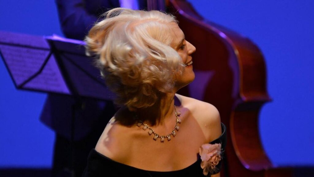 A woman in a black dress, adorned with a floral accessory and necklace, smiles while performing on stage. A double bass is seen in the background.