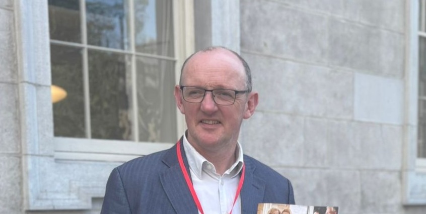 Johnny Sheehan in a blue suit and glasses stands outside holding a magazine. He has a red lanyard around his neck. The mood is casual and professional.
