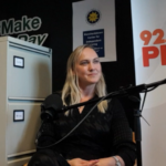 A woman is seated in a radio studio with a microphone in front of her. Behind her are filing cabinets, a "#Make Day" sign, and a "92.5 PHOENIX FM" banner. The atmosphere is professional and engaged.