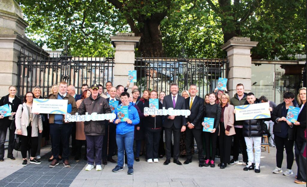 A diverse group of people holds banners and signs reading "Rehab Group" and advocacy messages. They stand in front of an iron fence under tree shade, conveying unity and determination.