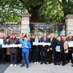 A diverse group of people holds banners and signs reading "Rehab Group" and advocacy messages. They stand in front of an iron fence under tree shade, conveying unity and determination.
