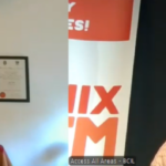 Two men are in a video call. The man on the left is in a room with certificates on the wall, wearing glasses. The man on the right wears headphones and a sweater, with a banner in the background. Both appear focused.