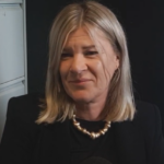 A woman with shoulder-length blonde hair smiles softly, wearing a black top and pearl necklace. She sits in an office setting with a cabinet and banner.