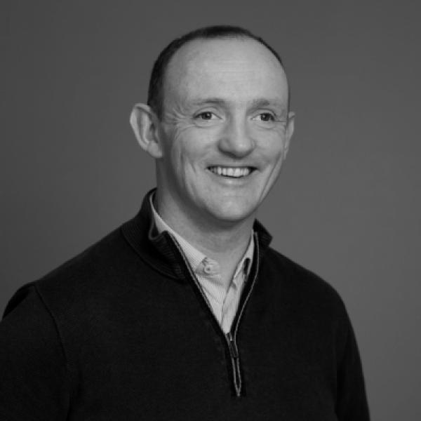 Johnny Sheehan, Member Engagement Manager with The Wheel - a man with short hair smiles warmly, wearing a dark zip sweater over a collared shirt. The black-and-white image has a neutral background.