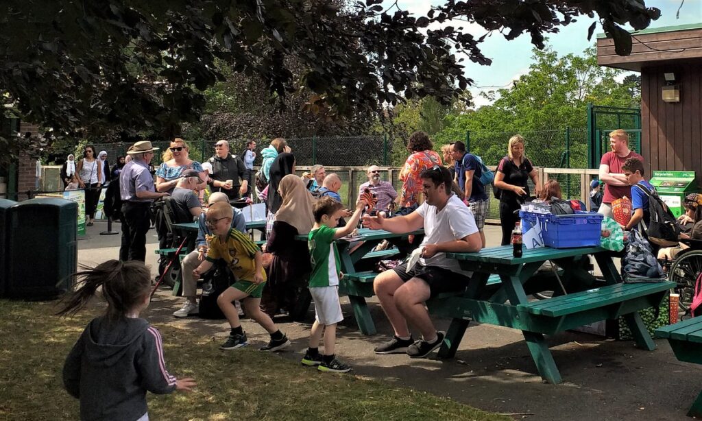 Outdoor gathering with people of various ages around picnic tables under trees. Children play nearby, while adults converse and relax. Bright, sunny atmosphere.
