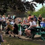 Outdoor gathering with people of various ages around picnic tables under trees. Children play nearby, while adults converse and relax. Bright, sunny atmosphere.